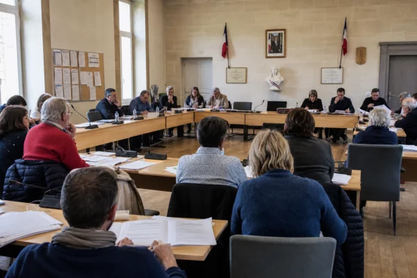 Salle municipale française lors d’un débat politique, avec élus anonymes, micros, dossiers et une chaise vide