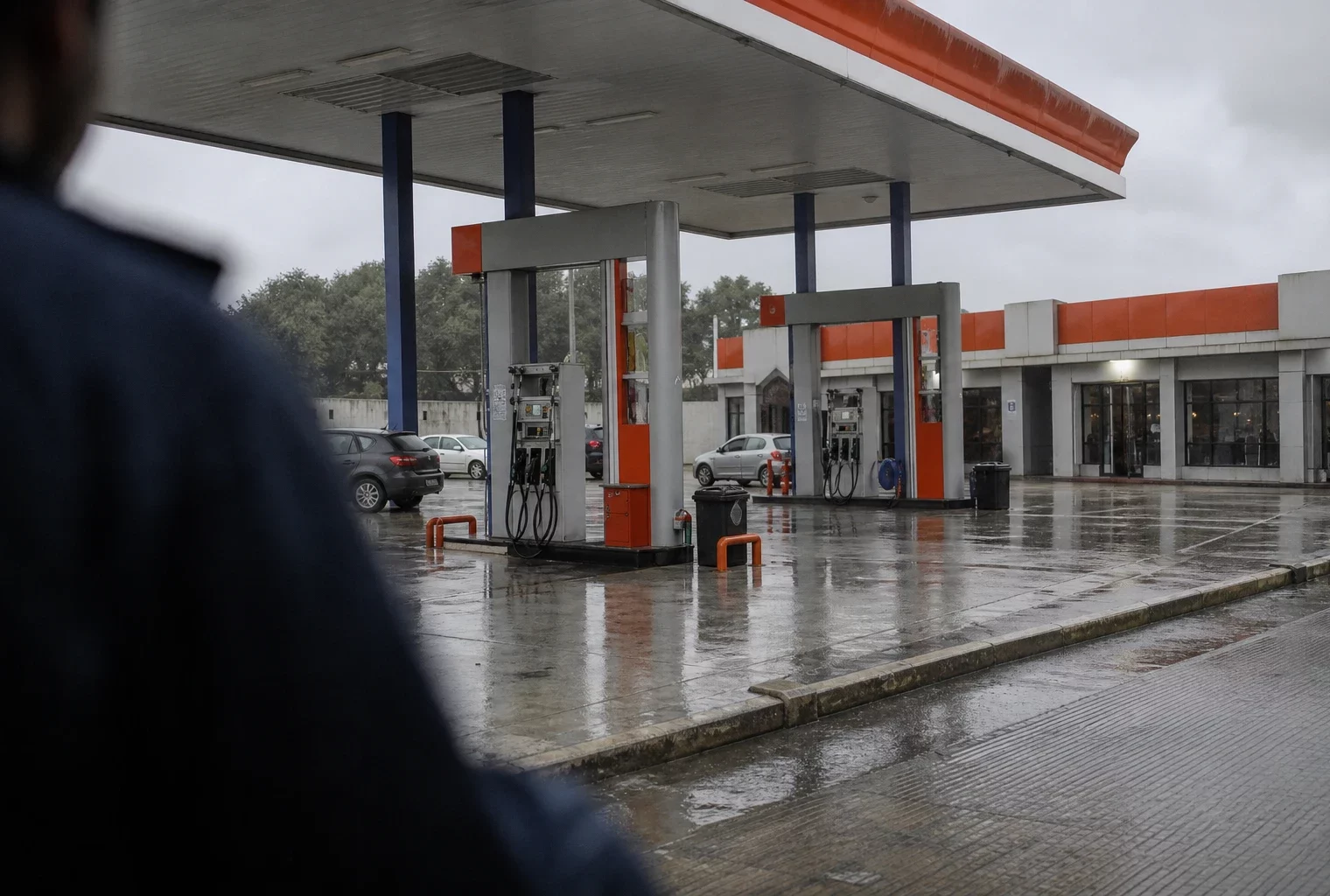 Station-service française sous la pluie, pompes à carburant et véhicules en attente, reflets humides sur l’aire vide.