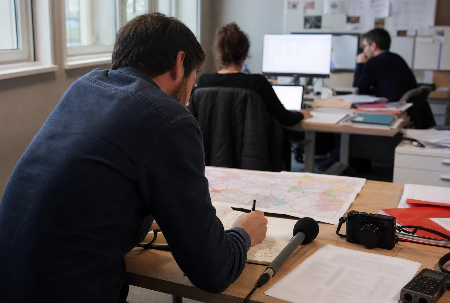 Journaliste préparant un sujet sur les maires dans une rédaction locale française, avec carnet et carte floue