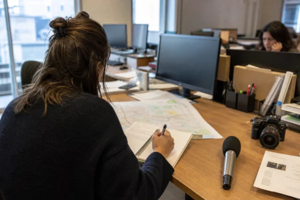 Journaliste préparant un sujet local dans une rédaction française, avec carnet, carte floue, micro et caméra de presse