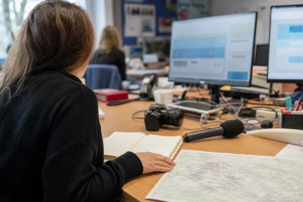 Journaliste préparant un reportage électoral dans une rédaction régionale, avec carnet, micro et carte floue sur le bureau