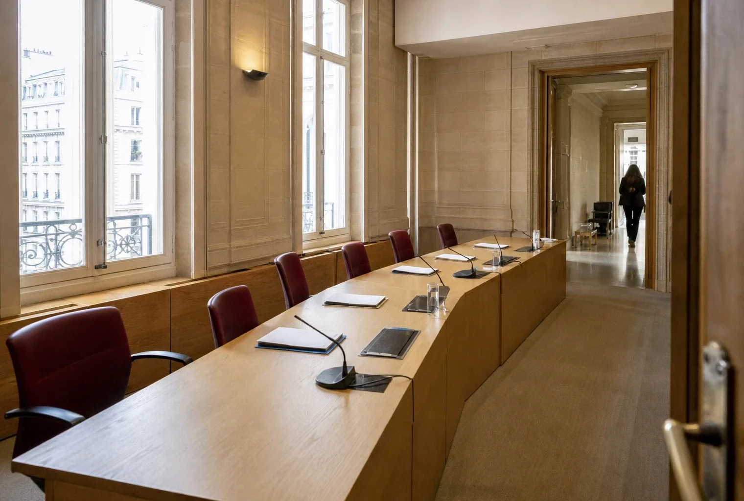 Salle d’audition claire d’une institution judiciaire française, avec micros, dossiers anonymes et couloir officiel en arrière-plan