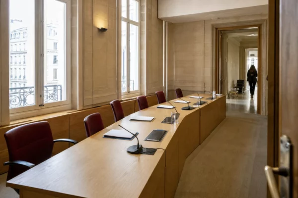 Salle d’audition claire d’une institution judiciaire française, avec micros, dossiers anonymes et couloir officiel en arrière-plan