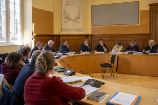 Salle de conseil municipal française avec une chaise vide, des micros et des dossiers lors d’une réunion politique