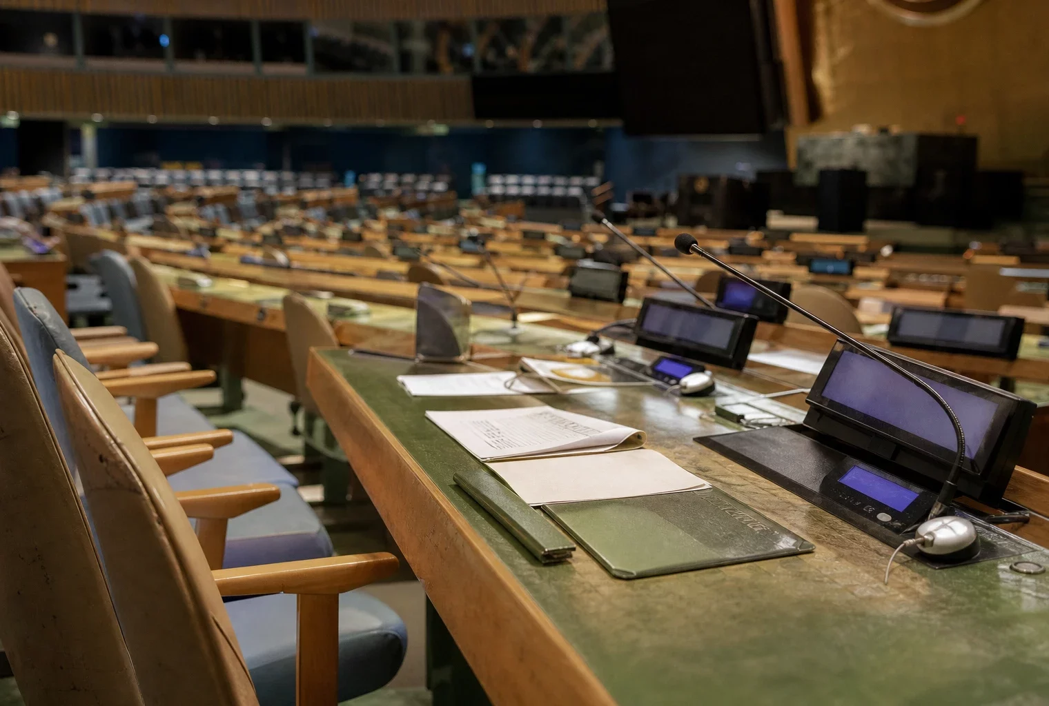 Salle de l’Assemblée générale de l’ONU à New York, rangées de pupitres, micros et dossiers sous une lumière diffuse.