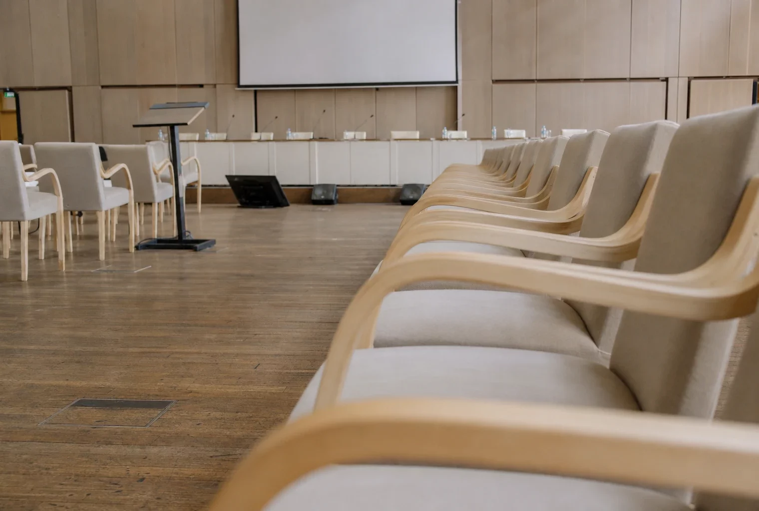 Salle de conférence vide avec rangées de chaises, pupitre et micros, lumière du matin et panneaux neutres en arrière-plan.