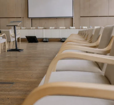 Salle de conférence vide avec rangées de chaises, pupitre et micros, lumière du matin et panneaux neutres en arrière-plan.