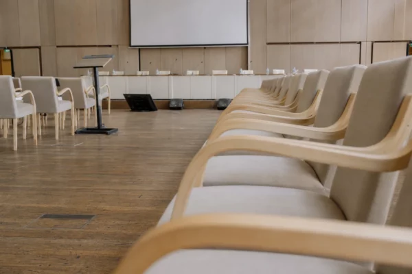 Salle de conférence vide avec rangées de chaises, pupitre et micros, lumière du matin et panneaux neutres en arrière-plan.