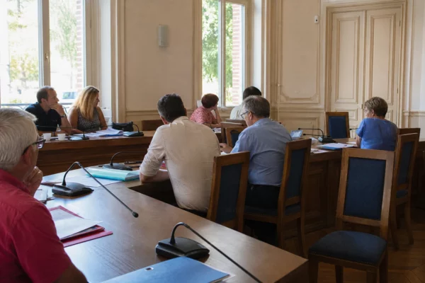 Salle de conseil municipal française avec élus anonymes, micros et une chaise vide autour d’une table en bois