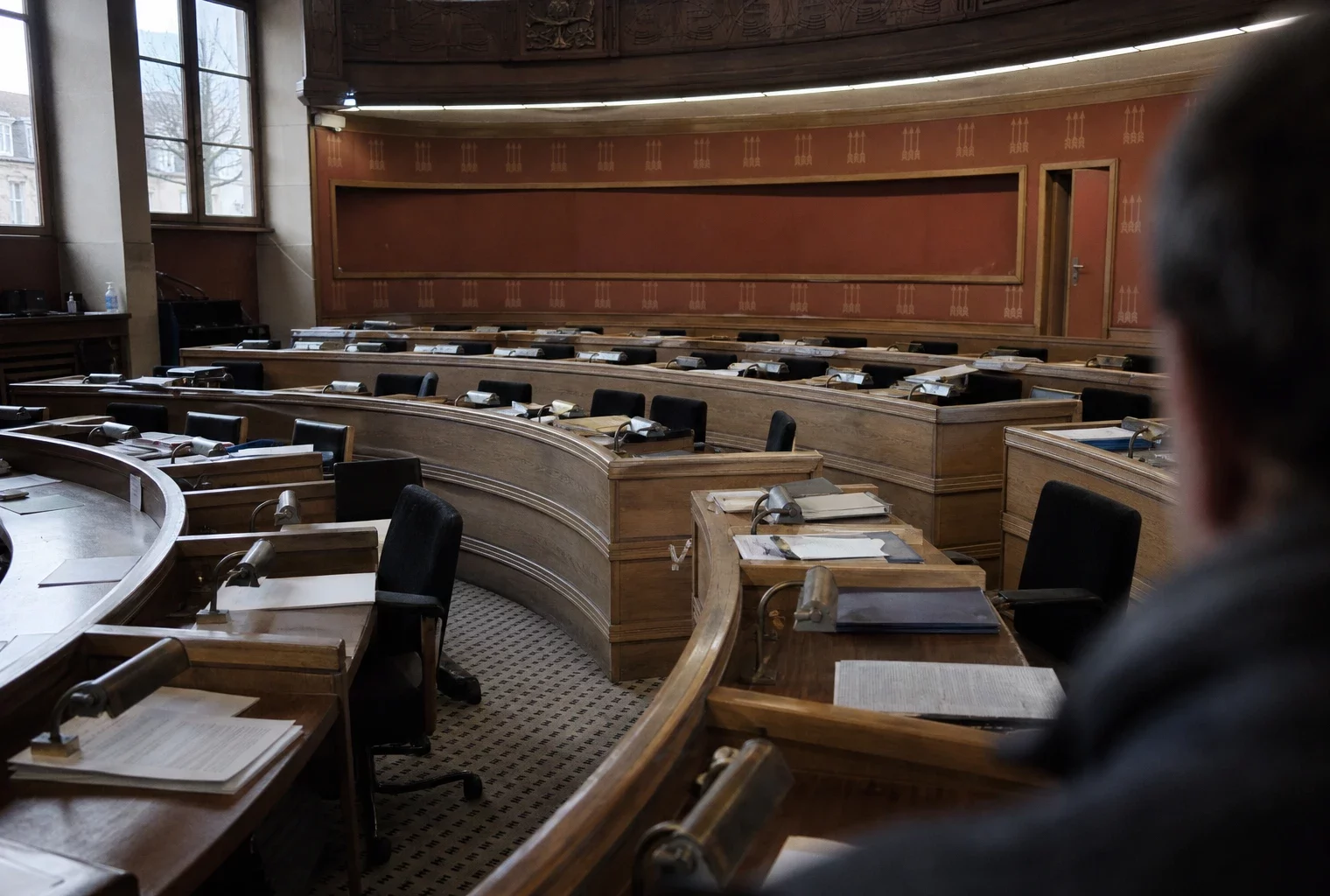 Salle du conseil municipal vide, tables en U et micros, lumière grise de pluie, ambiance sobre et humide.