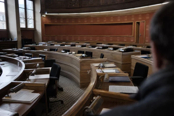 Salle du conseil municipal vide, tables en U et micros, lumière grise de pluie, ambiance sobre et humide.