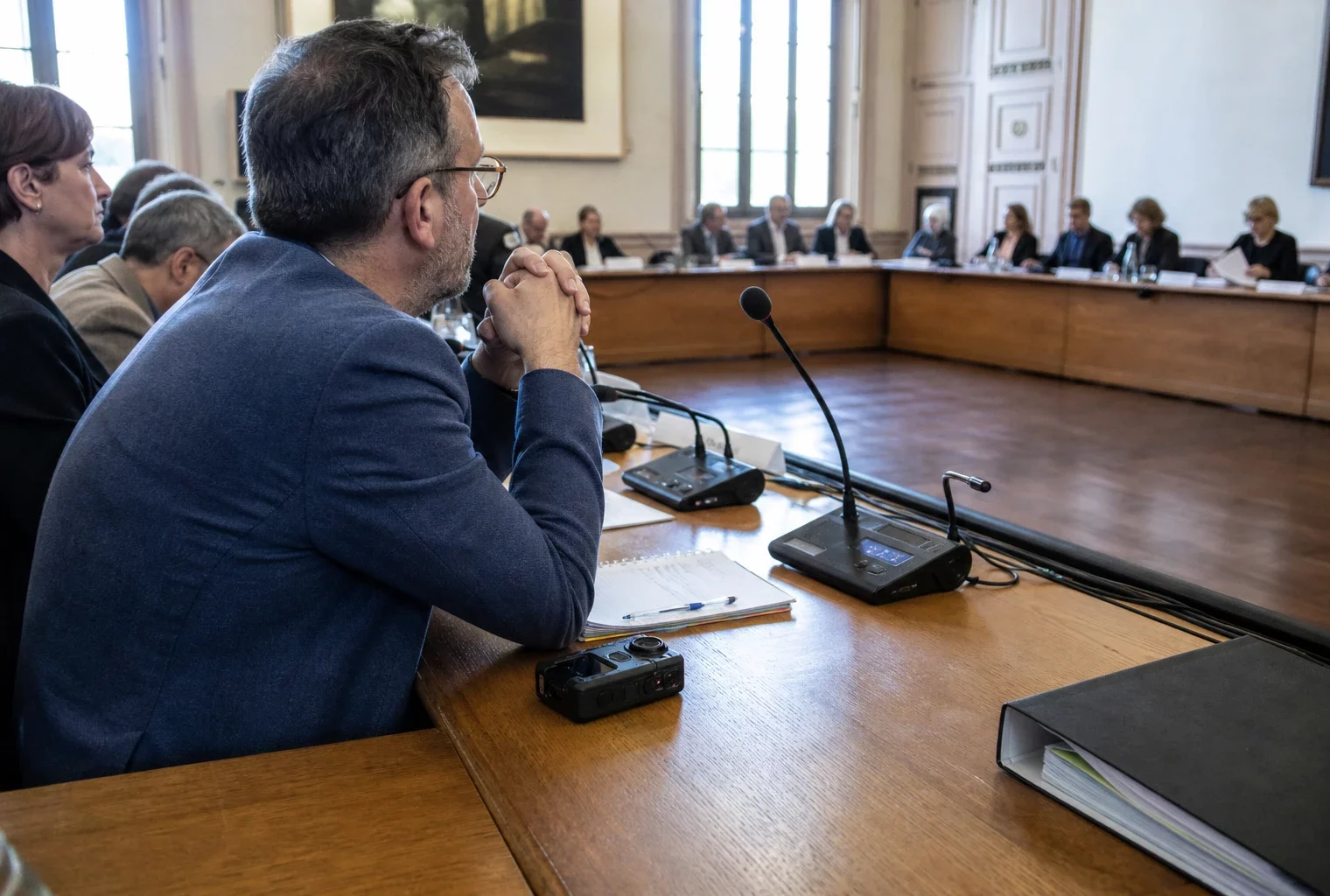 Salle municipale française avec chaise vide, micros et dossiers flous, évoquant une enquête sur la police municipale
