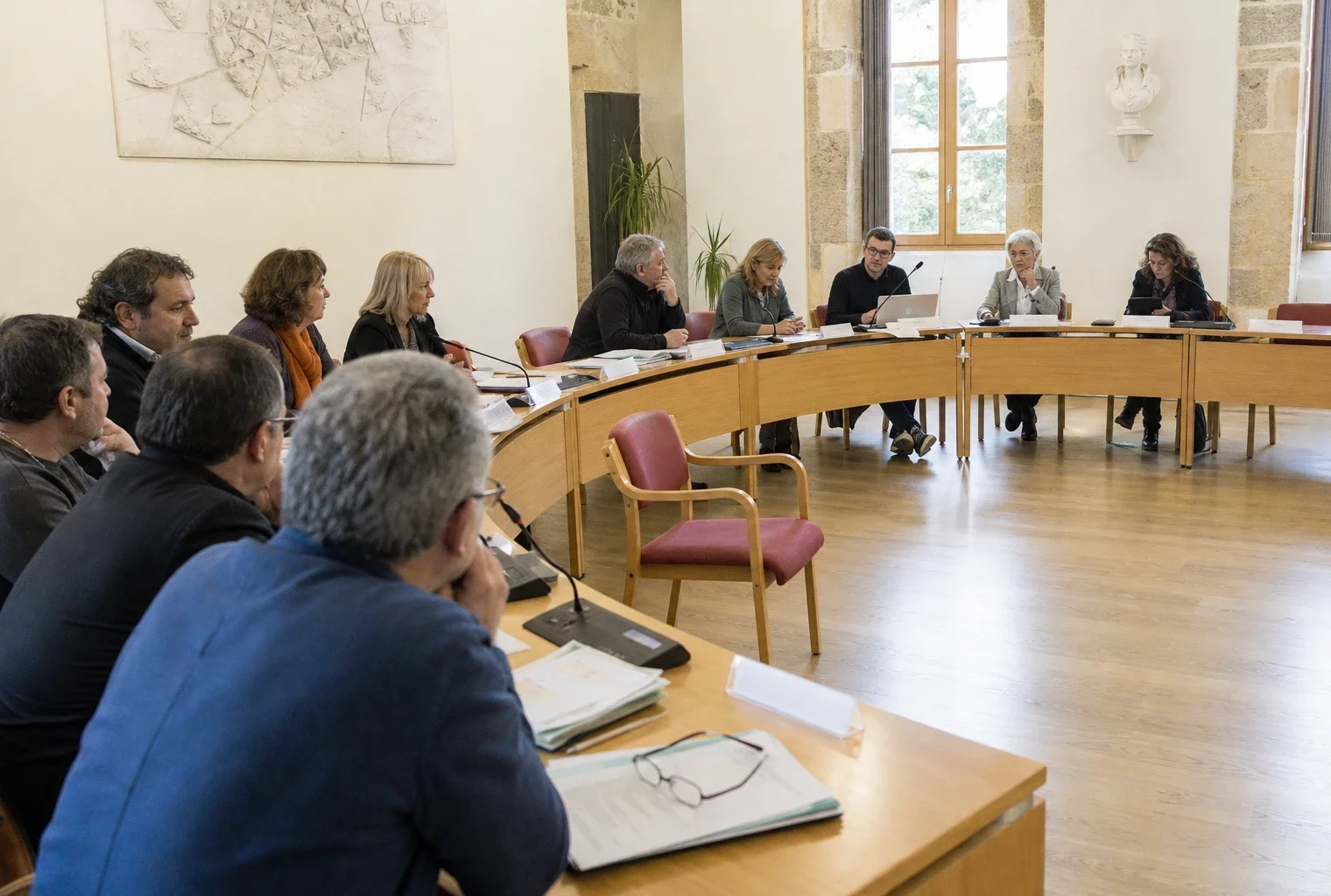 Salle de conseil municipal française avec une chaise vide, des micros et des élus anonymes en discussion