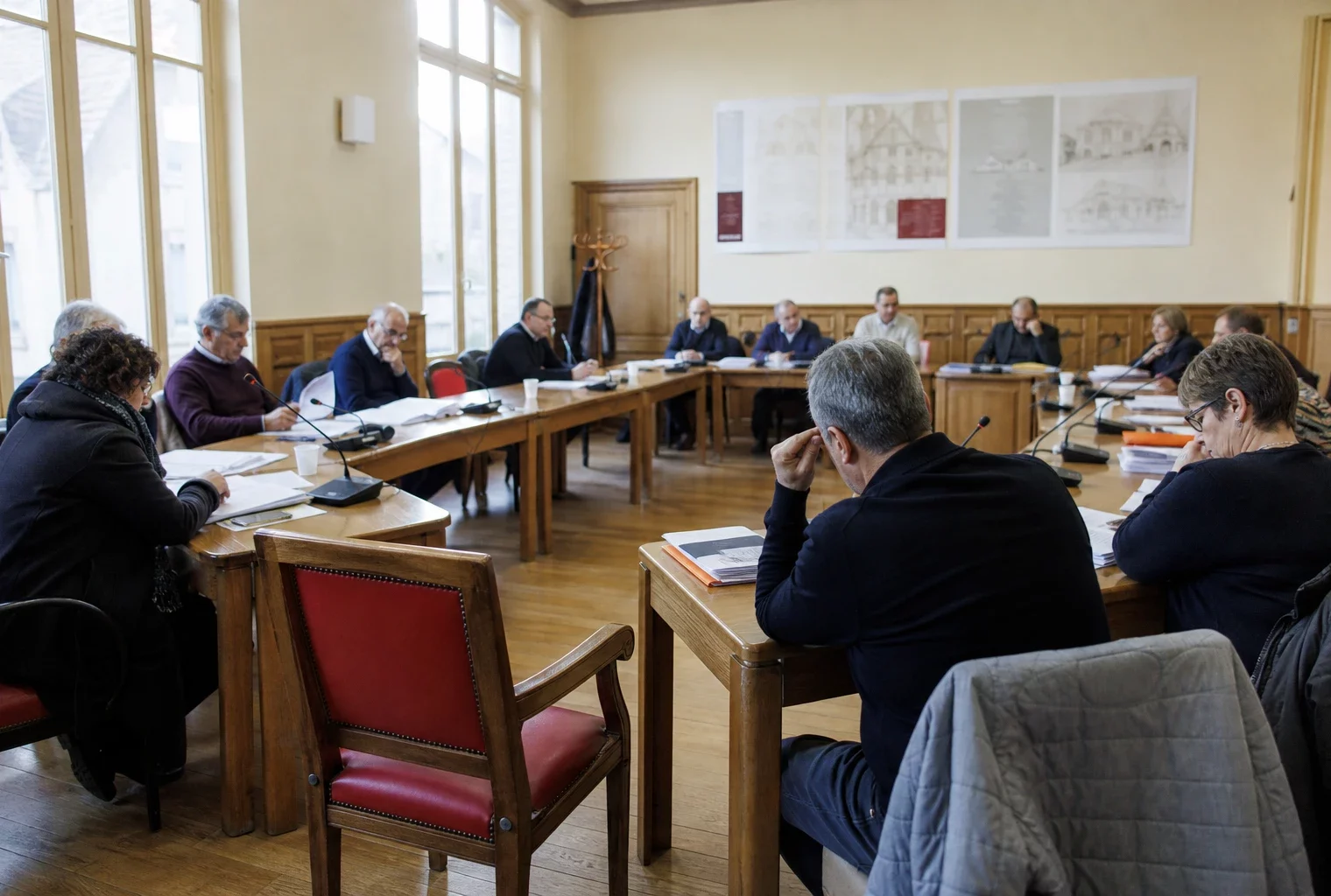 Salle de conseil municipal française avec élus anonymes, micros, dossiers et une chaise vide en vue oblique