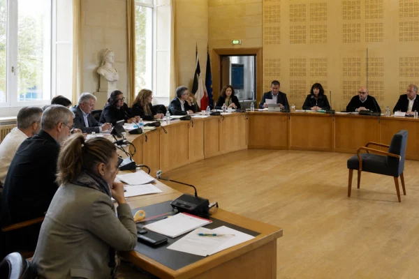 Salle de conseil municipal française avec élus locaux, micros, dossiers flous et une chaise vide en marge de la réunion