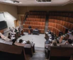 Amphithéâtre universitaire en bois avec pupitres et micros au centre, rangées de sièges vides et mur de fond sobre.