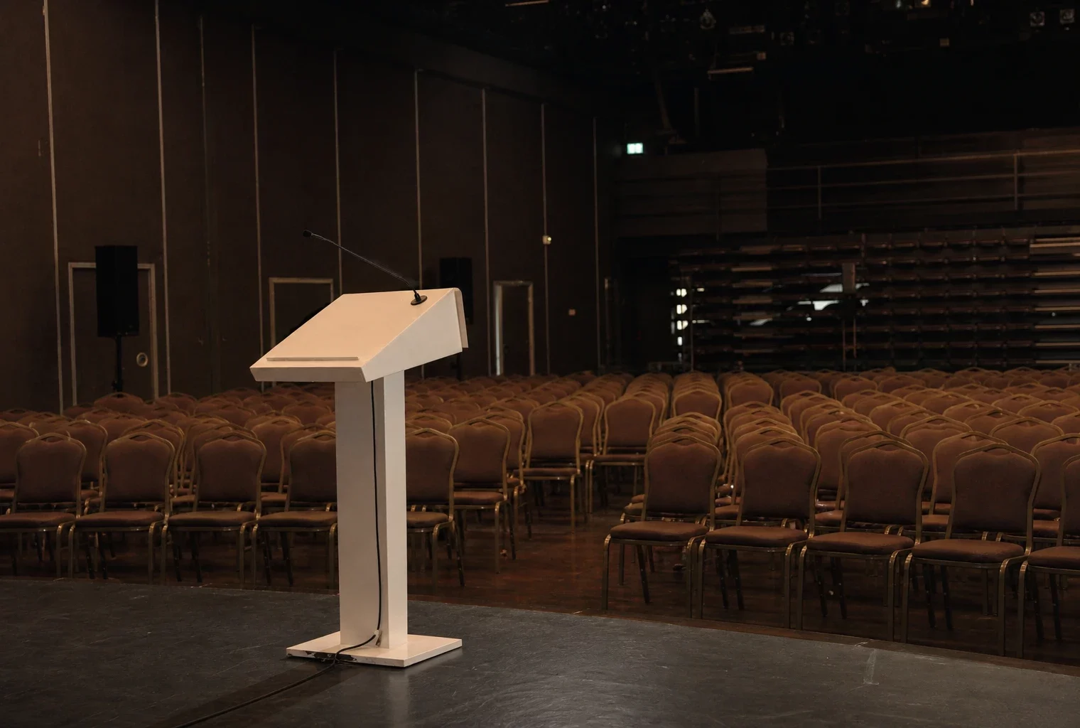 Salle de meeting vide à Bercy, rangées de chaises face à un pupitre blanc sous éclairage de contre-jour.