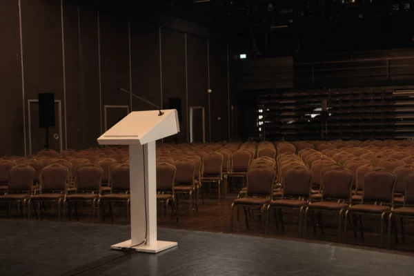 Salle de meeting vide à Bercy, rangées de chaises face à un pupitre blanc sous éclairage de contre-jour.