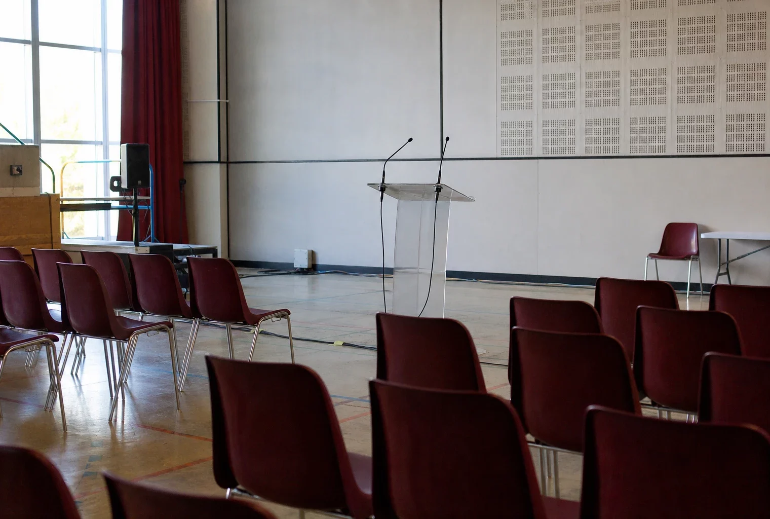 Salle de meeting de campagne vide avec rangées de chaises, pupitre et micros dans une salle polyvalente.