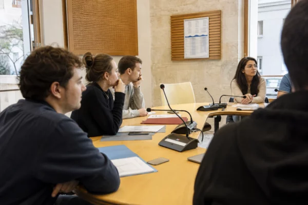 Jeunes volontaires réunis dans une salle municipale autour de micros, avec une chaise vide au premier plan
