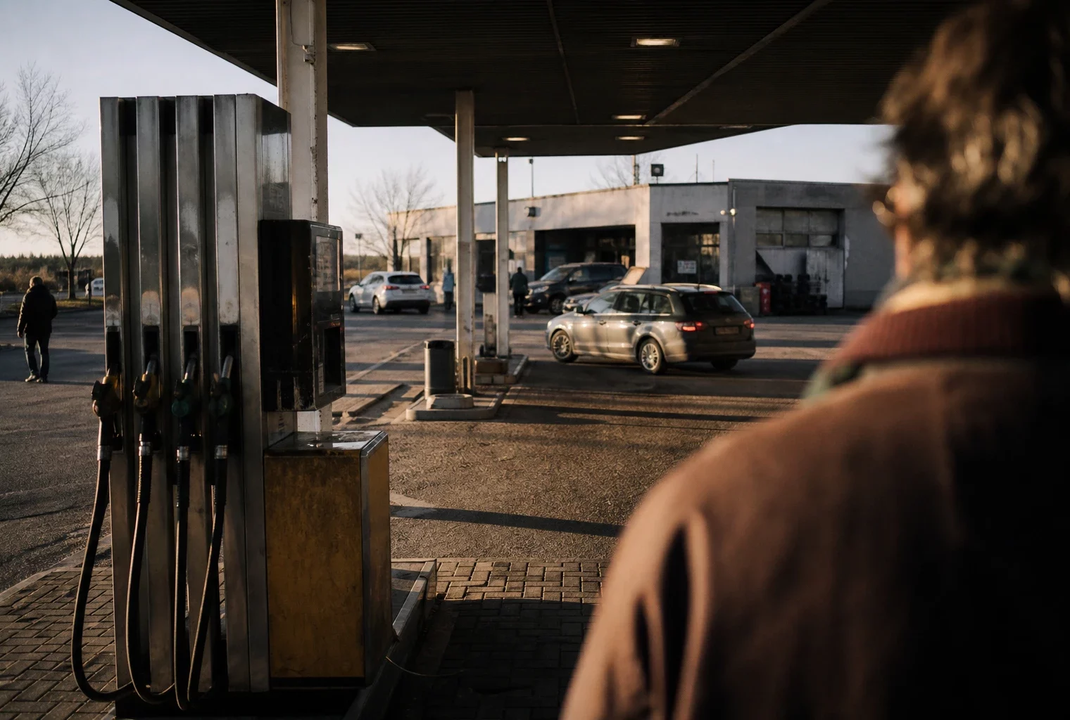 Station-service en périphérie, pompes usées au premier plan et voitures de passage sous une lumière d’hiver rasante.