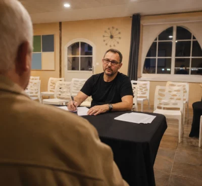 Salle de réunion municipale à Grenay, avec un élu assis à table portant un tee-shirt à slogan visible.