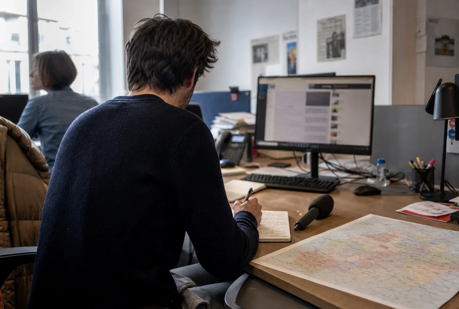 Journaliste préparant un sujet politique dans une rédaction locale française, avec carnet, carte floue et micro sans logo