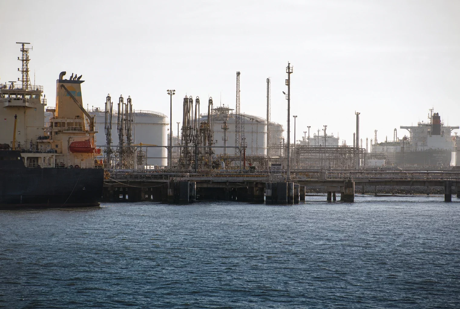Terminal pétrolier saoudien en activité, cuves, tuyauteries et bras de chargement devant des tankers et quais industriels.