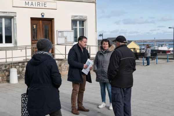 Habitants et élus anonymes échangent devant une mairie de Saint-Pierre-et-Miquelon, dans une lumière claire de bord de mer.