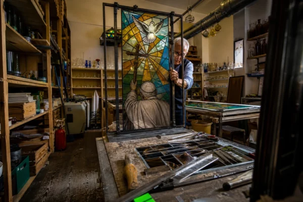 Dans un atelier de verrier en France, un grand vitrail contemporain se monte parmi outils, châssis et tables encombrées.
