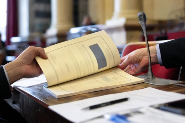 Mains anonymes feuilletant un dossier lors d’une audition parlementaire, avec micro et documents flous