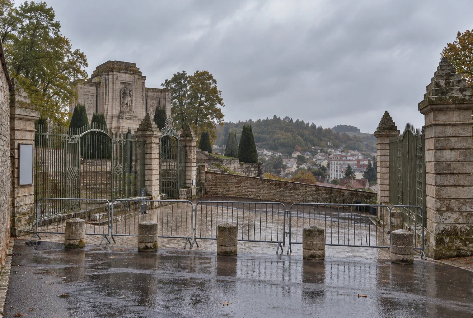 Des barrières et des bornes de pierre contrôlent l’entrée fermée d’un monument public, devant une façade de pierre sous la pluie.