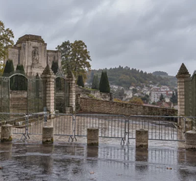 Des barrières et des bornes de pierre contrôlent l’entrée fermée d’un monument public, devant une façade de pierre sous la pluie.