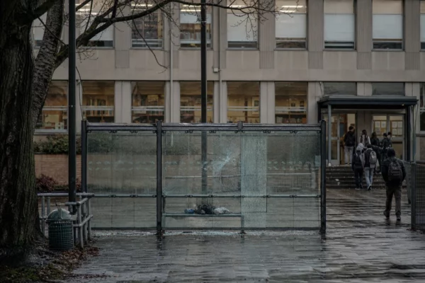Abribus dégradé devant un lycée, vitre fendue, banc vide et trottoir humide sous une lumière froide.
