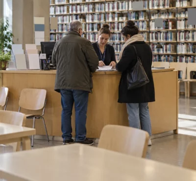 Dans le hall d’une bibliothèque, un comptoir temporaire oriente des usagers parmi des chaises, des formulaires et des rayonnages.