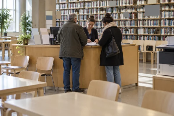 Dans le hall d’une bibliothèque, un comptoir temporaire oriente des usagers parmi des chaises, des formulaires et des rayonnages.
