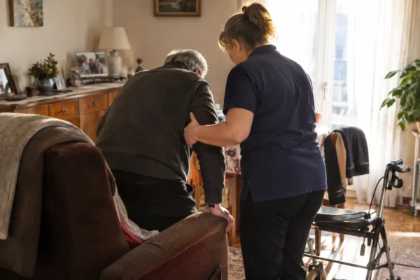 Dans son salon, un homme âgé se lève de son fauteuil avec l'aide d'une auxiliaire de vie, près de son déambulateur.