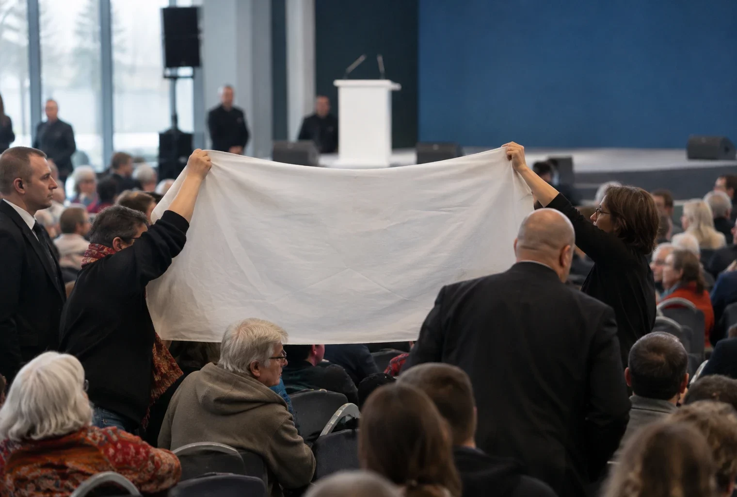 Une banderole unie est tenue entre les rangées de chaises d’une salle de meeting, près d’agents de sécurité.
