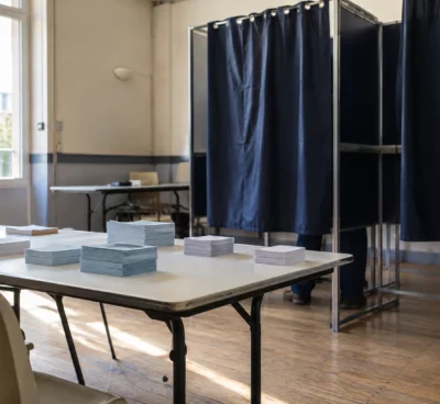 Des bulletins vierges sont posés sur une table devant les isoloirs d’une salle de vote municipale.