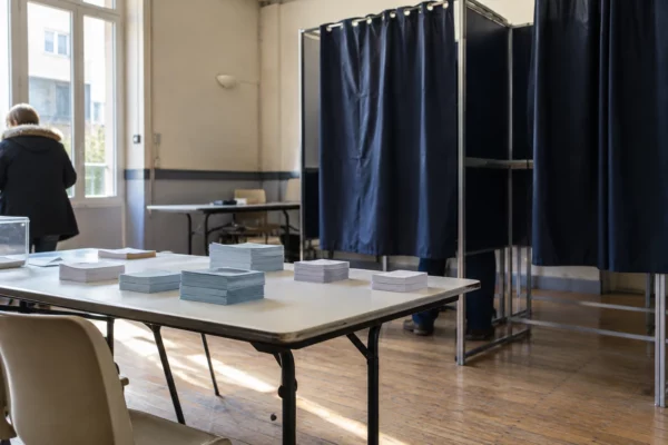 Des bulletins vierges sont posés sur une table devant les isoloirs d’une salle de vote municipale.
