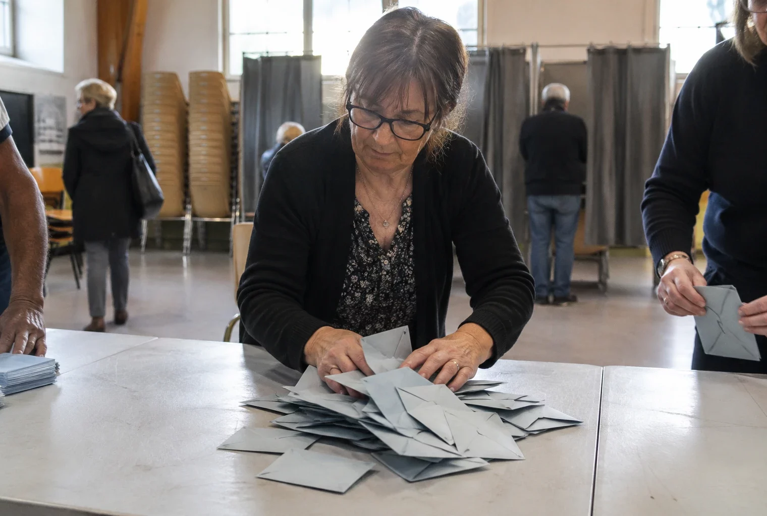 Des bulletins pliés et des enveloppes sont rassemblés sur une table, avec des isoloirs et des chaises en arrière-plan.