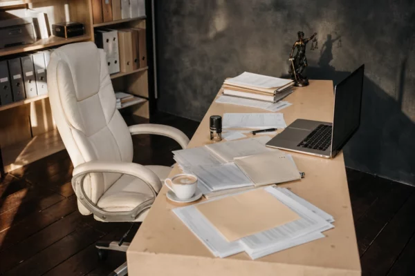 Bureau de commissaire de justice vu de haut, avec ordinateur ouvert, dossiers imprimés, tampon et étagère de classeurs.