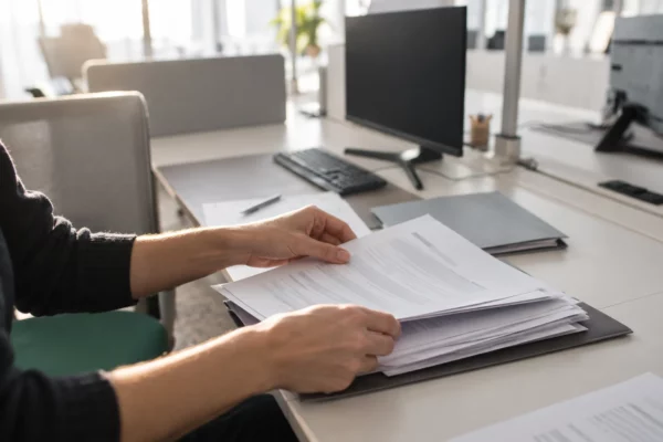 Des mains anonymes trient des dossiers administratifs sur un bureau partagé, parmi des écrans éteints et du matériel de travail.