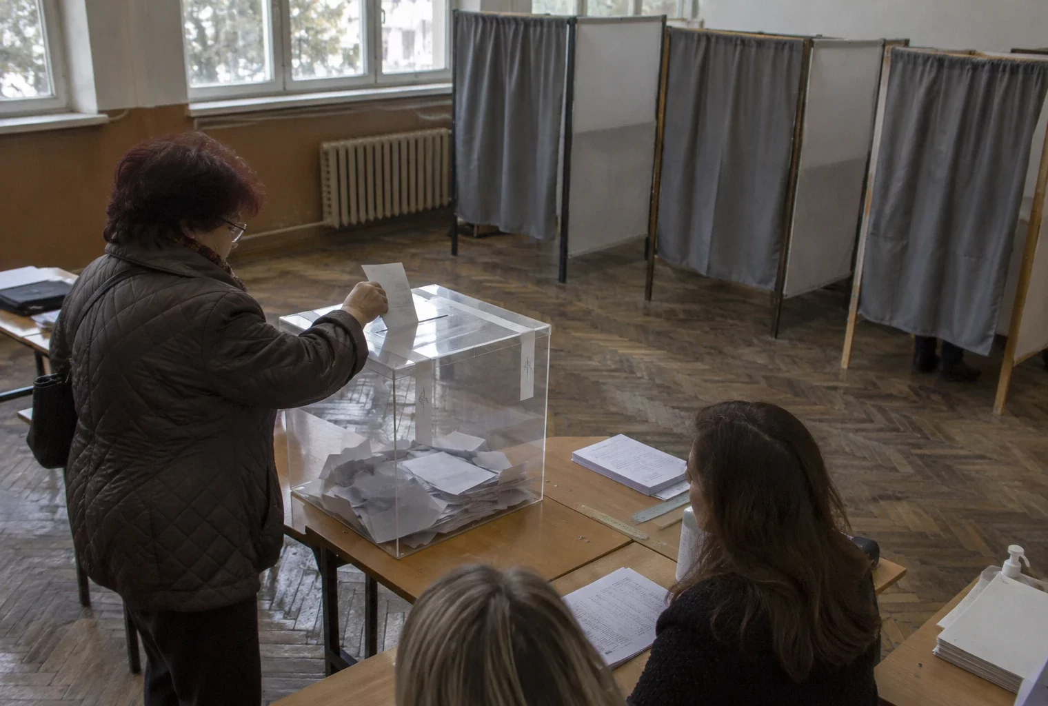 Urne électorale et isoloirs dans un bureau de vote sobre, avec quelques bulletins visibles sur une table.