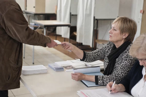 Une salle de vote installée avec isoloirs, table d’émargement et bulletins, image d’une primaire en préparation.