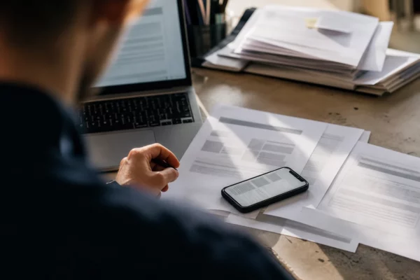 Bureau de travail avec smartphone, feuilles de conversations imprimées, ordinateur ouvert et dossiers empilés en arrière-plan.