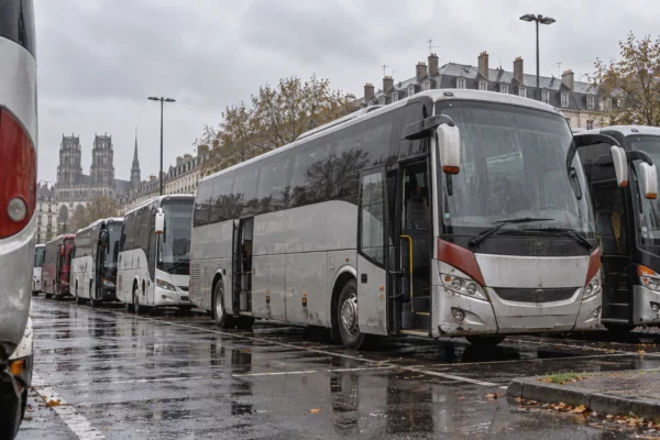 Des autocars stationnés sur une aire urbaine mouillée, entre marquages au sol effacés et bâtiments ordinaires.
