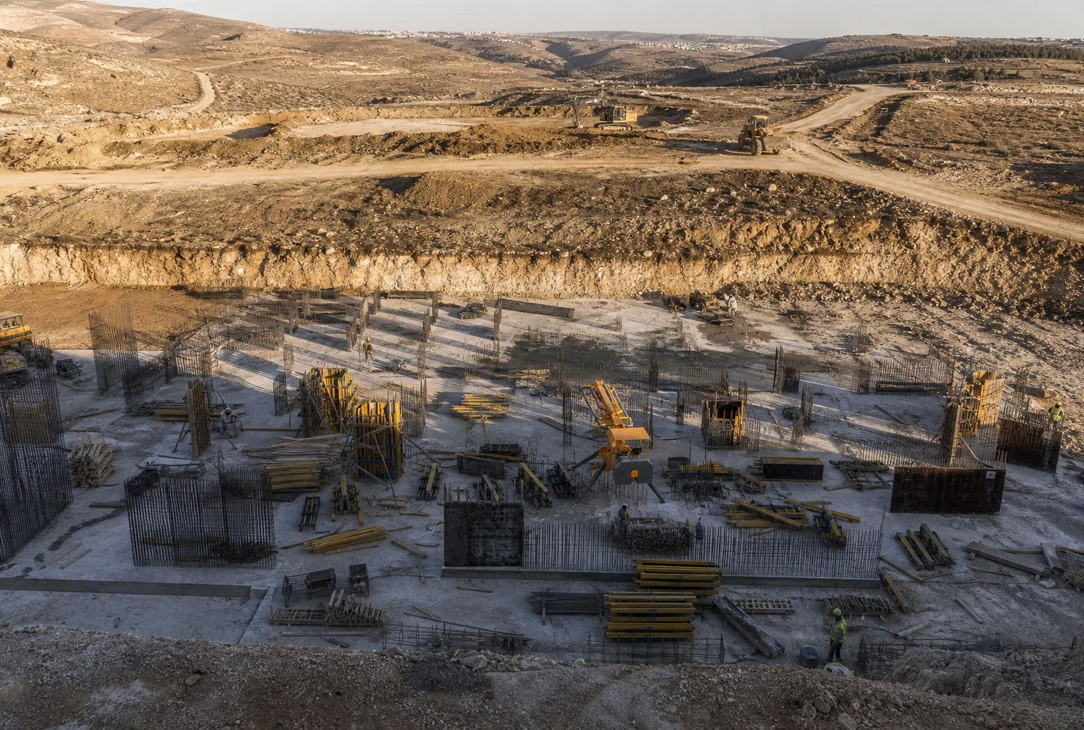 Vue plongeante d’un chantier E1 en Cisjordanie, avec terrassements, engins, talus de terre et collines basses au fond.