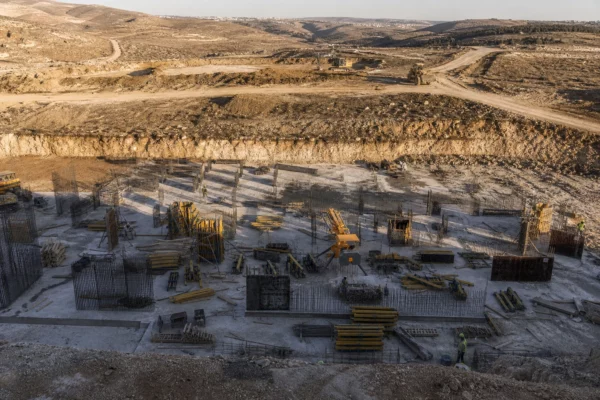 Vue plongeante d’un chantier E1 en Cisjordanie, avec terrassements, engins, talus de terre et collines basses au fond.