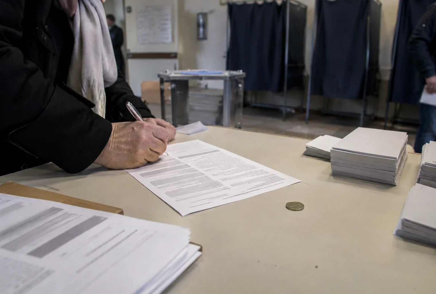 Une charte papier, un stylo et une pièce d’un euro sur une table, devant des isoloirs et des bulletins vierges.